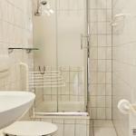 Badezimmer mit Dusche, Waschbecken und Toilette. Weiße Fliesen und Handlauf.