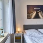 Schlafzimmer mit Bett, Nachttisch und Fenster. Wandbild zeigt Brücke mit Schriftzug.