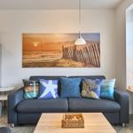 Ein Wohnzimmer mit Sofa, Kissen und Wandbild. Fenster mit Vorhängen und Tischlampe.