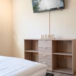 Schlafzimmer mit Bett, Schrank und Wandfernseher. Auf dem Schrank steht ein HOME-Schild.