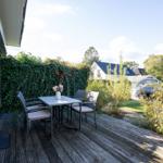 Terrasse mit Tisch und vier Stühlen, umgeben von grüner Hecke und Pflanzen.