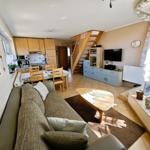 Wohnzimmer mit Sofa, Esstisch und Küchenbereich. Holztreppe führt nach oben.