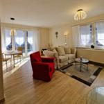 Living room with sofa, red armchair, and dining area. Wooden floor and large windows.
