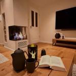 Wohnzimmer mit Kamin, Fernseher und Holzboden. Auf Tisch: Buch, Teekanne und Tassen.