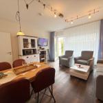 Esszimmer mit Tisch und Stühlen, Wohnbereich mit Sofa und Sesseln, TV und Fenster mit Vorhängen.