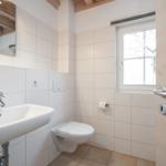 Bathroom with sink, toilet, and window. White tiles and wooden beams on ceiling.