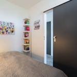 Schlafzimmer mit Bett, Wandbild und Bücherregal. Tür führt in einen Flur.