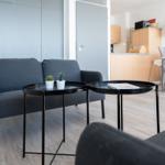 Ein Wohnbereich mit grauem Sofa, zwei schwarzen Tischen und Pflanzen. Im Hintergrund Küche und Essbereich.