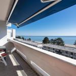 Terrasse mit Blick auf Meer und Strand. Blauer Sonnenschutz und weiße Gartenstühle.