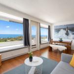 Apartment with sea view, dining table, and sofa. Large windows and wall art with lighthouse.