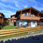 Wooden house with balcony, blue shutters, and garden fence. Backyard with terrace and chairs.