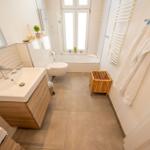 Badezimmer mit Badewanne, Waschbecken und Toilette. Holzregal und Handtücher hängen an der Wand.
