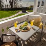 Terrasse mit Tisch, zwei Gläsern und Trauben. Blick auf Garten und Wasser.