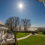 Haus mit Terrasse, Garten und Blick auf Wasser unter sonnigem Himmel.