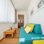 Ein Wohnzimmer mit blauem Sofa, gelben Kissen und Holztisch. Durchgang zur Küche.