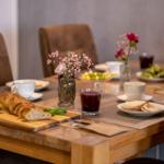 Ein gedeckter Holztisch mit Brot, Obst und Getränken. Blumenarrangements und Teller sind sichtbar.