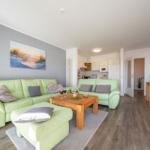 Wohnbereich mit grünen Sofas, Holztisch und Küchenbereich im Hintergrund.