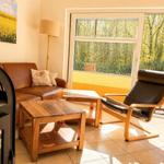 Living area with leather sofa, wooden table, and chair. Entrance to terrace with view into the forest.