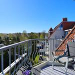 Balkon mit Stuhl und Tisch, Blick auf Dächer und Bäume unter blauem Himmel.
