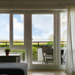 Durch das Fenster blickt man auf einen Balkon mit Stuhl und Blick auf das Meer.
