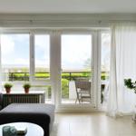 Wohnzimmer mit Blick auf Balkon und Grünfläche. Vorhänge, Sofa, Pflanzen und Tisch sind sichtbar.