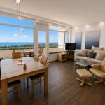 Living and dining area with view of water and greenery. Wooden table, sofa, and chair.