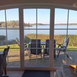 Large window view of a lake. Terrace with table and chairs. Indoor area with dining table and sofa.