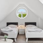 Two beds under sloped ceilings with round window. Wooden floor and gray seating area.