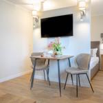 Ein Raum mit Tisch, zwei Stühlen, Fernseher und Bett im Hintergrund. Blumen und Holzboden.
