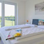 Schlafzimmer mit Balkon, Blick auf Wiese und Wald, Bett mit Kissen und Decke.
