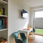 Ein Raum mit Schreibtisch, Fernseher und Bücherregal. Durchgangstür mit Jalousie zum Garten.