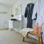 Badezimmer mit Dusche, Toilette und Waschbecken. Davor hängen Bademäntel und steht ein Holzwaschbecken.