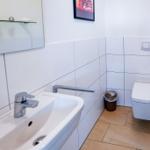 Bad mit Waschbecken, Spiegel, Toilette und Mosaikfliesen an den Wänden.