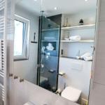 Badezimmer mit Dusche, Spiegelschrank und Heizkörper. Auf Regalen liegen Handtücher und Dekorationsobjekte.