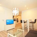 Living area with TV, glass table, and bar stools. Kitchen and bedroom are visible.
