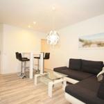 Living room with sofa, glass table, and bar stools. Wall art and ceiling light visible.