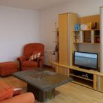 Living room with orange sofa, armchair, and wooden furniture. TV shows sea view.