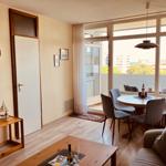 Living area with dining space, balcony, and wooden floor. Windows overlook the city.