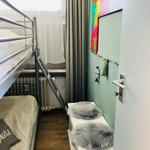 Zimmer mit Doppelstockbett, Fenster, Wandbild und grauen Fellkissen auf einem weißen Regal.