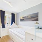 Schlafzimmer mit Bett, Schreibtisch und Wandbild. Fenster mit blauen Vorhängen.