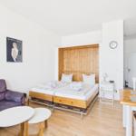 Room with two beds, sofa, table, and wall clock. Wooden surfaces and white walls.