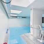 Indoor pool with white tiled walls and glass partition. Wooden steps and handrail.