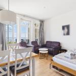 A room with a bed, sofa, table and chairs. Through windows you can see the sea.