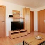 Wohnzimmer mit Fernseher, Holzmöbeln und Tisch. Mehrere Türen und Wanddekoration sichtbar.