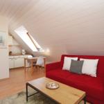 Ein Wohnbereich mit rotem Sofa, Holztisch und schrägem Dach. Im Hintergrund Küche und Tisch.