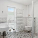 Badezimmer mit Dusche, Waschbecken und Toilette. Weiße Fliesen und gemusterte Bodenfliesen.