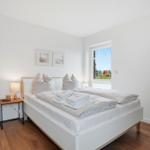 Schlafzimmer mit Bett, Nachttisch und Fenster. Spiegel und Holzboden sichtbar.
