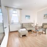 Living room with sofa, armchair, glass table, and wooden floor. Windows with white curtains.