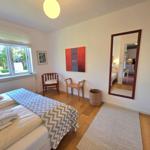 Schlafzimmer mit Bett, Fenster, zwei Stühlen und Spiegel. Holzboden und Wandbild.