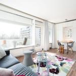 Living area with sofa, glass table, and floral rug. Dining area with table and chairs. Kitchen and balcony visible.